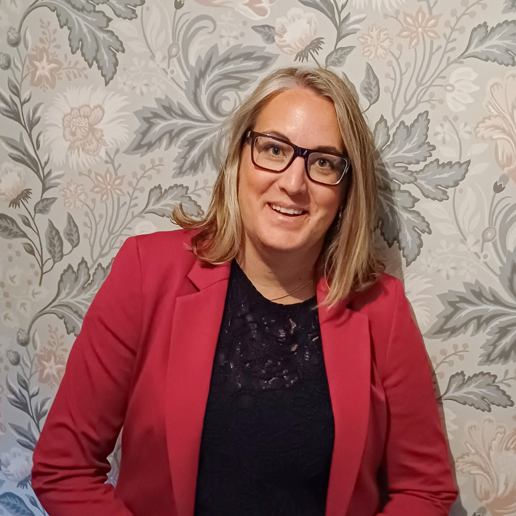 Om Starlings Arkitekt Malin Starenflykt ler. Hon har på sig en röd kavaj och svarta glasögon. I bakgrunden är en blommigt mönstrad tapet.